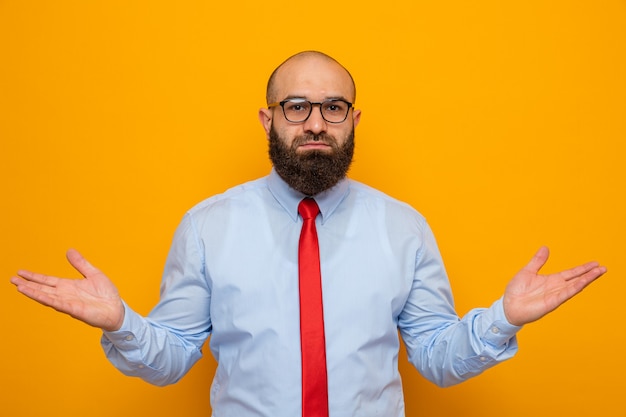 Free photo bearded man in red tie and shirt wearing glasses looking confused spreading amrs to the sides