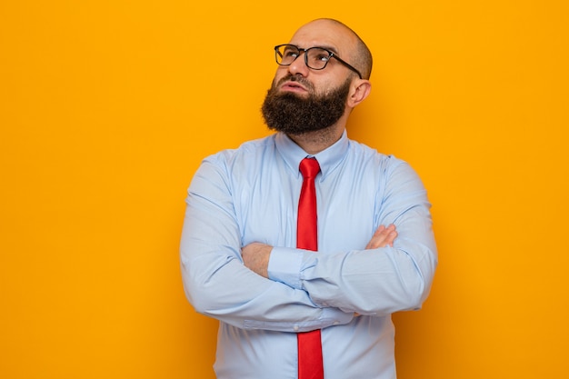 Free photo bearded man in red tie and shirt wearing glasses looking up with pensive expression with arms crossed