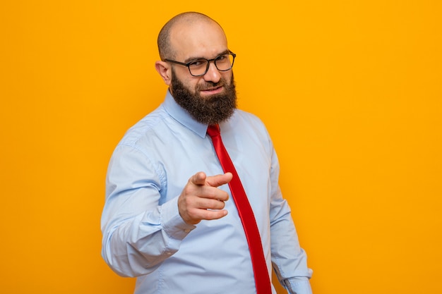 Bearded man in red tie and shirt wearing glasses pointing with index finger at front, smiling cheerfully looking