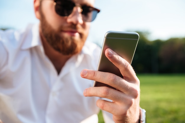 Бородатый мужчина в солнцезащитные очки и рубашку при использовании смартфона. Фокус на телефоне