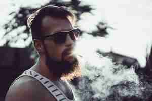 Free photo a bearded man in sunglasses smokes a cigarette at sunset, releases a thick tobacco smoke