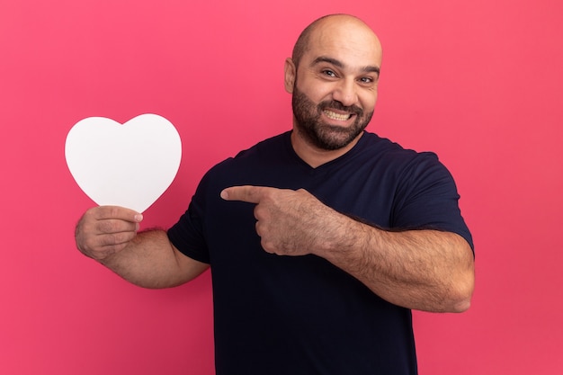 Uomo barbuto in maglietta che tiene il cuore del cartone che indica con il dito indice sorridendo con la faccia felice che sta sopra il muro rosa