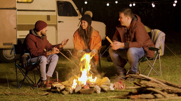 Bearded man telling a funny joke to his friends around camp fire. Retro camper van. Camping tent.