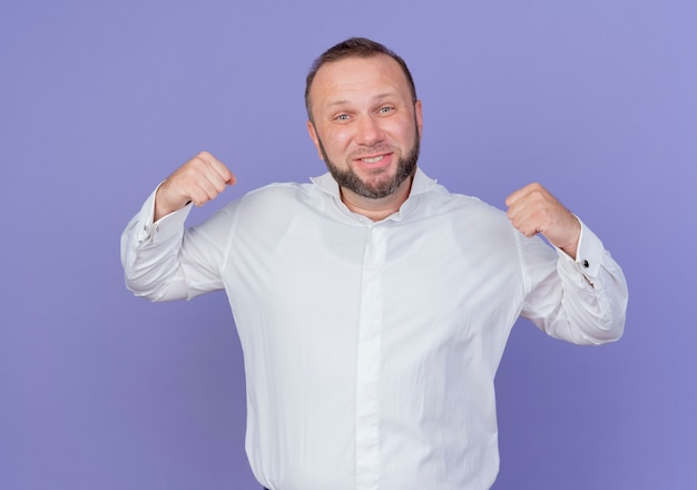 Foto gratuita uomo barbuto che indossa una camicia bianca stringendo i pugni felici ed entusiasti che gioiscono del suo successo in piedi sopra il muro blu