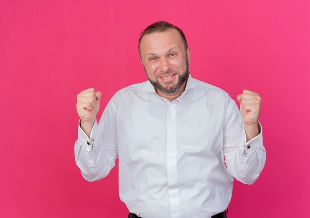 Бесплатное фото Бородатый мужчина в белой рубашке, сжимая кулаки, счастлив и взволнован, стоя у розовой стены