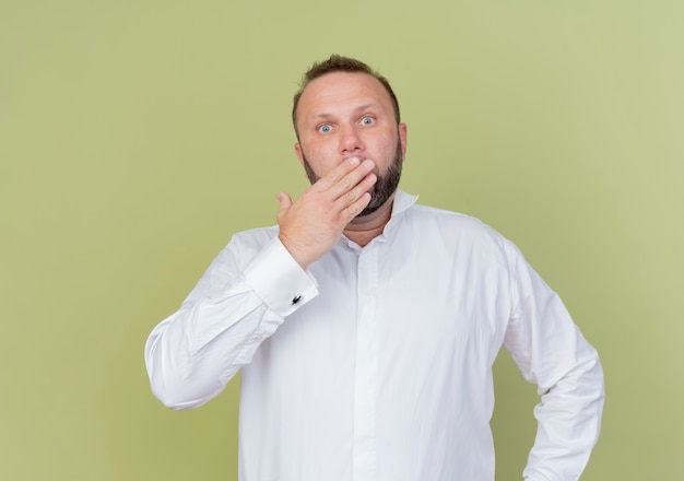 Foto gratuita uomo barbuto che indossa una camicia bianca che copre la bocca con la mano che è scioccato in piedi sopra la parete leggera