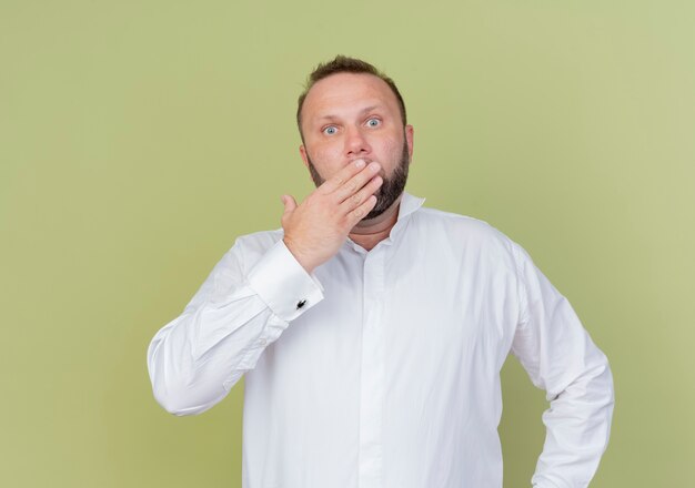 Bearded man wearing white shirt covering mouth with hand being shocked standing over light wall