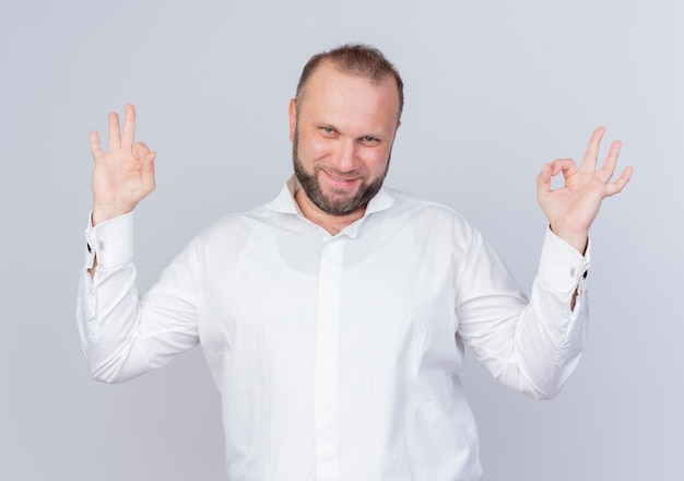 Bearded man wearing white shirt happy and positive smiling making ok sign standing over white wall