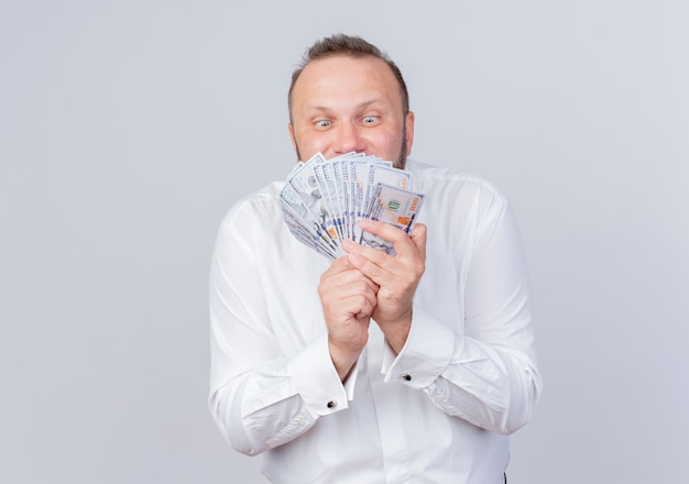 Bearded man wearing white shirt holding cash happy and excited standing over white wall