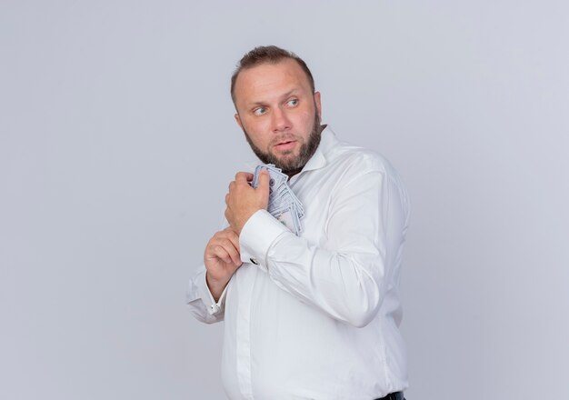 Bearded man wearing white shirt holding cash hiding money looking worried standing over white wall