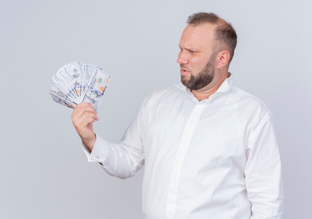 Bearded man wearing white shirt holding cash looking at money with skeptic expression standing over white wall