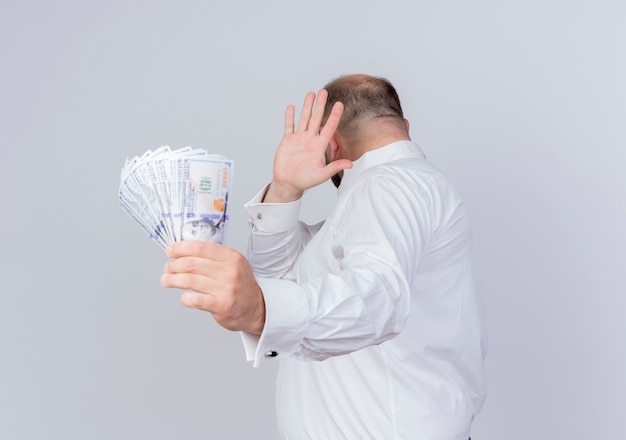Free photo bearded man wearing white shirt holding cash making defense gesture with hand against money standing over white wall