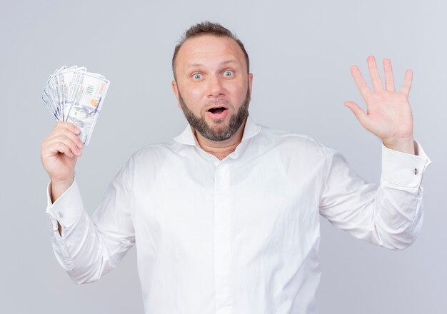 Bearded man wearing white shirt holding cash raising hand in surrender surprised and happy standing over white wall