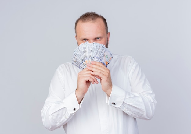Bearded man wearing white shirt holding cash  smiling slyly standing over white wall