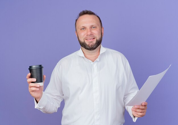 Bearded man wearing white shirt holding coffee cup and blank page looking  smiling standing over blue wall