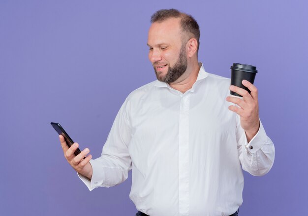 Bearded man wearing white shirt holding coffee cup looking at his smartphone screen smiling standing over blue wall