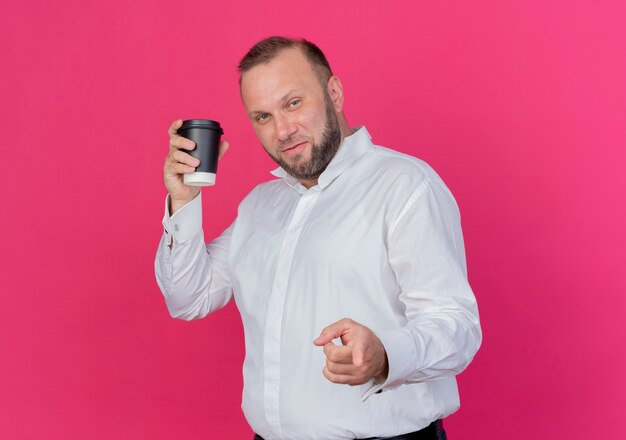 Bearded man wearing white shirt holding coffee cup pointign with index finger  smiling standing over pink wall