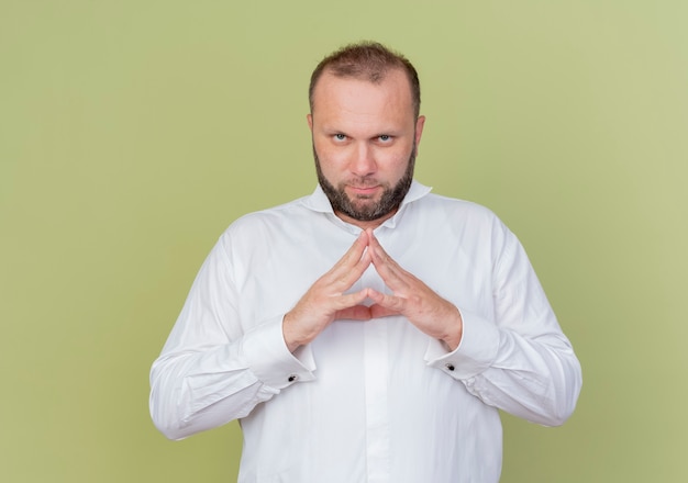 Foto gratuita uomo barbuto che indossa una camicia bianca tenendo le palme insieme in attesa con la faccia seria in piedi sopra la parete leggera