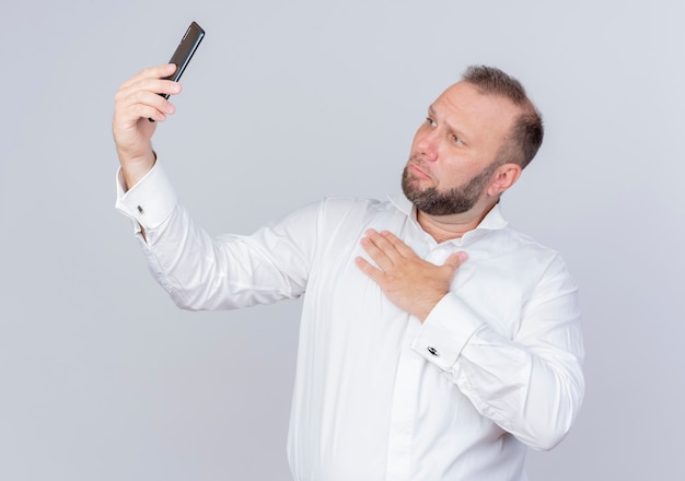 Бородатый мужчина в белой рубашке держит смартфон с видеозвонком, чувствуя благодарность за руку на груди, стоящую над белой стеной