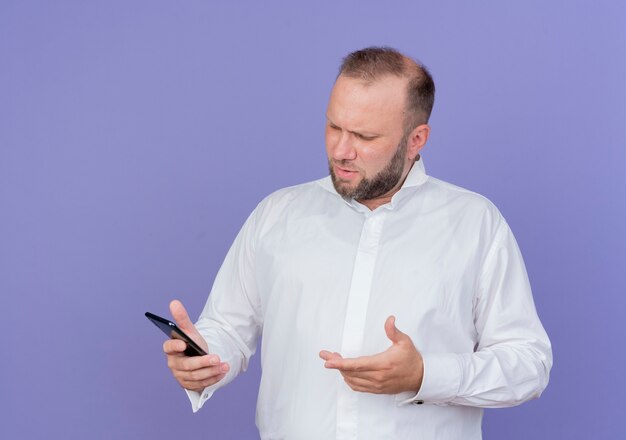 Bearded man wearing white shirt holding smartphone looking at screen confused standing over blue wall