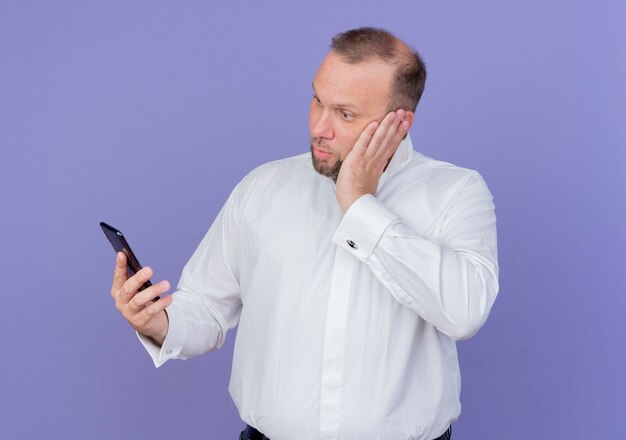 Bearded man wearing white shirt holding smartphone looking at screen with serious face being confused standing over blue wall