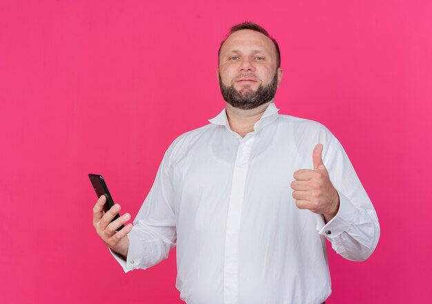 Bearded man wearing white shirt holding smartphone  showing thumbs up standing over pink wall