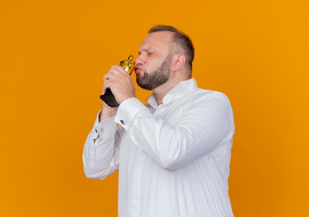 Bearded man wearing white shirt holding trophy kissing it happy and excited standing over orange wall