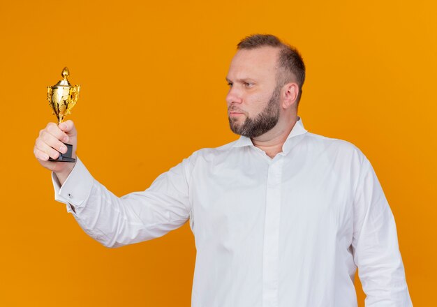 Bearded man wearing white shirt holding trophy looking at it with serious face standing over orange wall