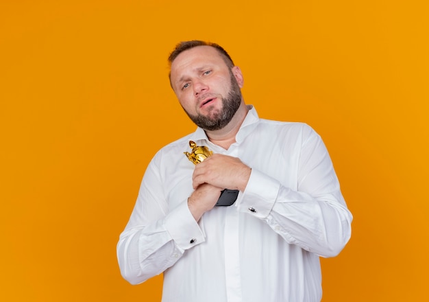 Bearded man wearing white shirt holding trophy with unhappy face standing over orange wall