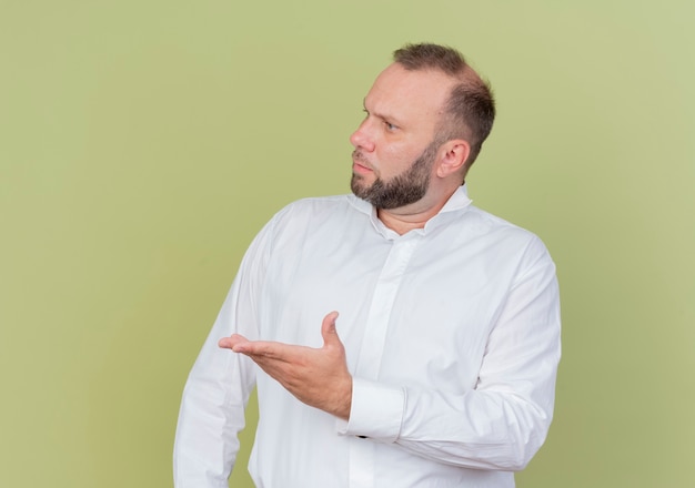 Bearded man wearing white shirt looking aside with arm out as asking displeased standing over light wall