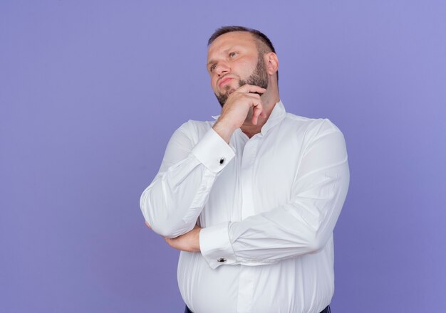Bearded man wearing white shirt looking aside with pensive expression with hand on chin thinking standing over blue wall