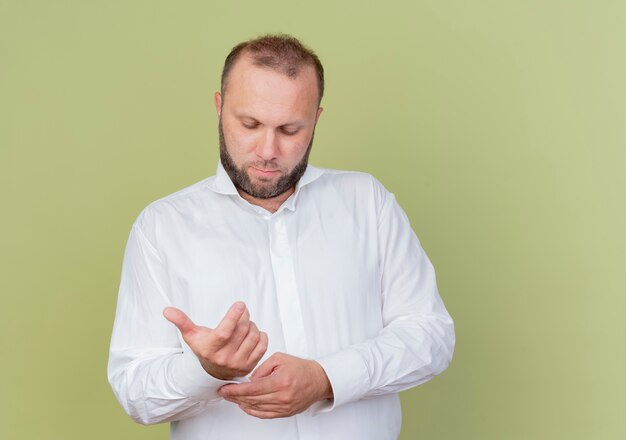 Bearded man wearing white shirt looking down fixing his cufflinks standing over light wall