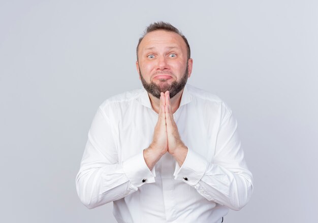 Bearded man wearing white shirt looking hands together like praying and begging with hope expression standing over white wall
