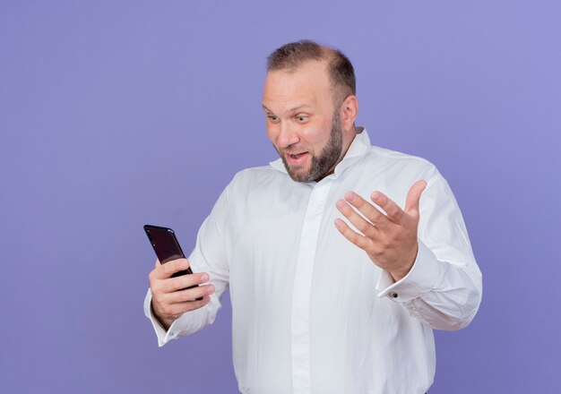 青い壁の上に立って幸せで興奮して笑顔の手で身振りで示す彼のスマートフォンの画面を見ている白いシャツを着たひげを生やした男