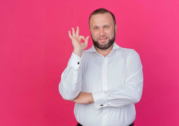 Bearded man wearing white shirt looking  smiling confident showing ok sign standing over pink wall