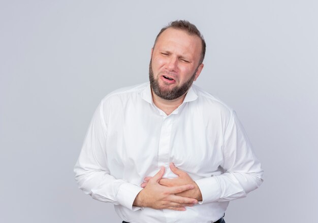 Bearded man wearing white shirt looking unwell touching his belly having pain standing over white wall