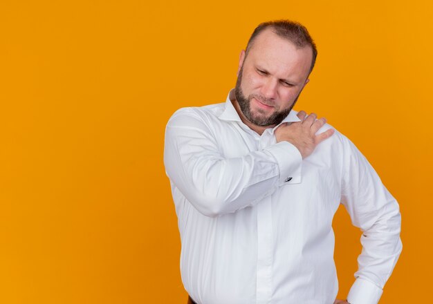 Bearded man wearing white shirt looking unwell touching his shoulder feeling pain standing over orange wall