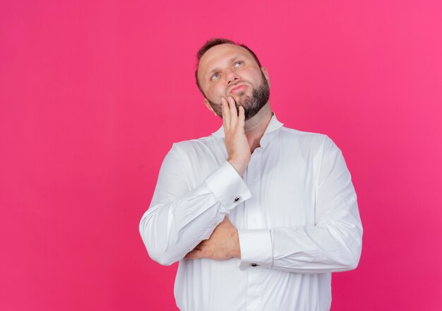 Bearded man wearing white shirt looking up with pensive expression on face thinking standing over pink wall