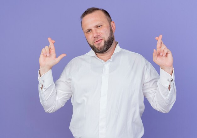 Bearded man wearing white shirt making desirable wish crossing fingers standing over blue wall