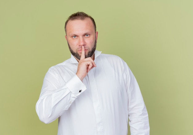 Бородатый мужчина в белой рубашке делает жест молчания с пальцем на губах, стоя над светлой стеной