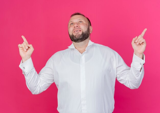 Uomo barbuto che indossa una camicia bianca rivolta verso l'alto con il dito indice sorridente felice e positivo in piedi oltre il muro rosa