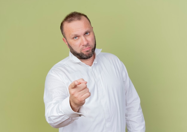 Free photo bearded man wearing white shirt pointing with index finger looking displeased standing over light wall