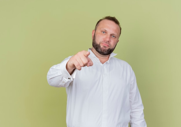 Bearded man wearing white shirt pointing with index finger standing over light wall