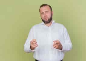 Free photo bearded man wearing white shirt pointing with index fingers looking confused standing over light wall
