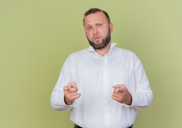Бородатый мужчина в белой рубашке, указывая указательными пальцами в замешательстве, стоит над светлой стеной