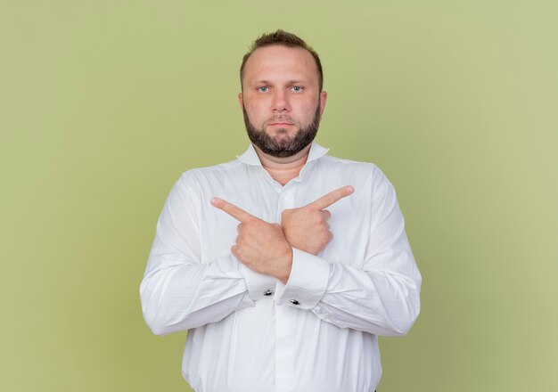 Bearded man wearing white shirt pointing with index fingers in opposite directions standing over light wall