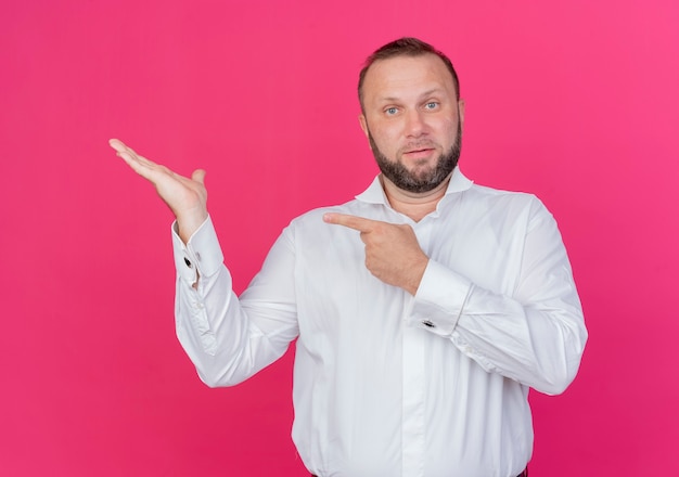 Bearded man wearing white shirt presenting something with arm pointing with finger to the side standing over pink wall