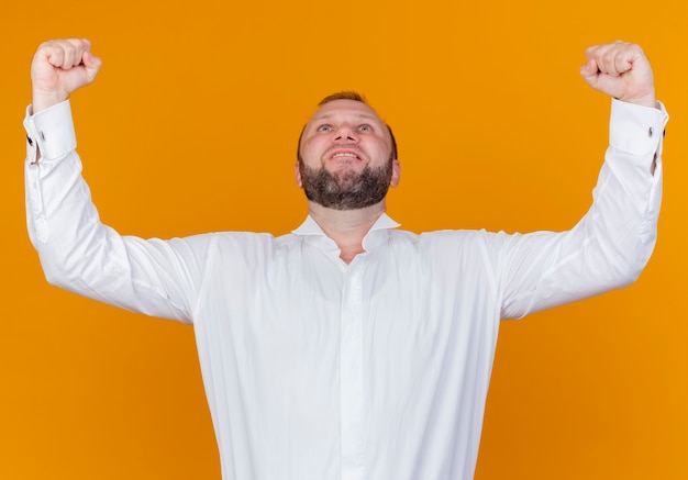 Foto gratuita uomo barbuto che indossa una camicia bianca alzando i pugni gioendo del suo successo in piedi sopra il muro arancione