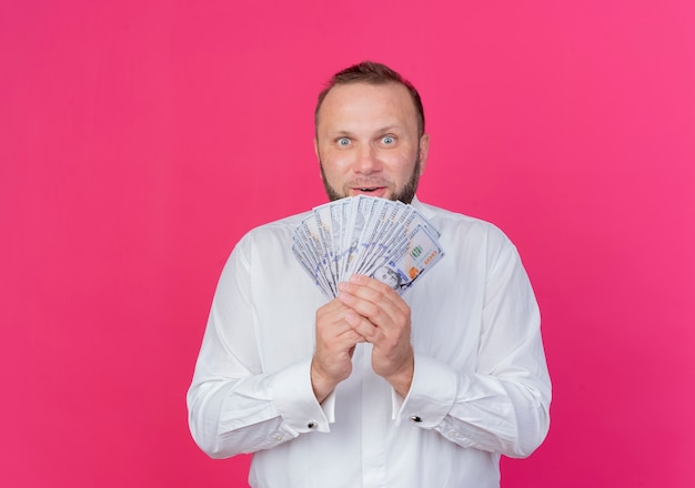 Foto gratuita uomo barbuto che indossa una camicia bianca che mostra contanti guardando sorpreso in piedi sopra la parete rosa