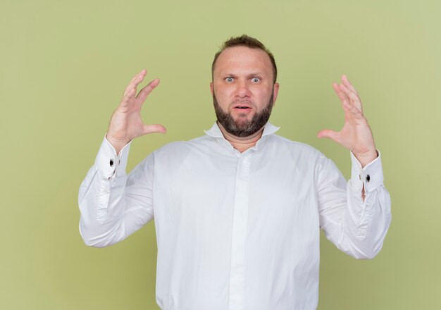 Bearded man wearing white shirt  showing large size gesture measure symbol standing over light wall